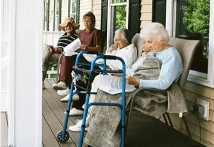 Four elderly individuals relaxing on a porch; one using a walker, another reading a newspaper.
