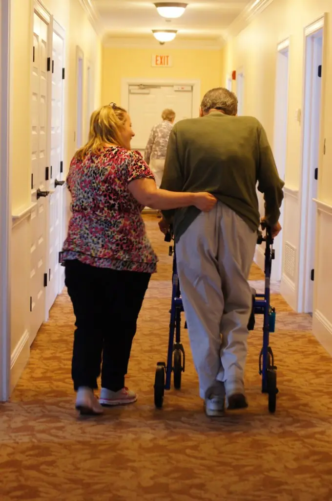 A woman assists an elderly man using a walker in a hallway, with another person further ahead.