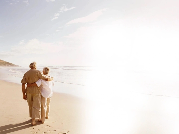 Elderly couple walking arm in arm along a sunny beach.