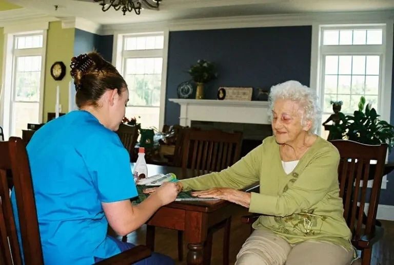 A nurse in blue scrubs holds the hands of an elderly woman at a wooden table in a brightly lit room, with a domestic setting in the background.